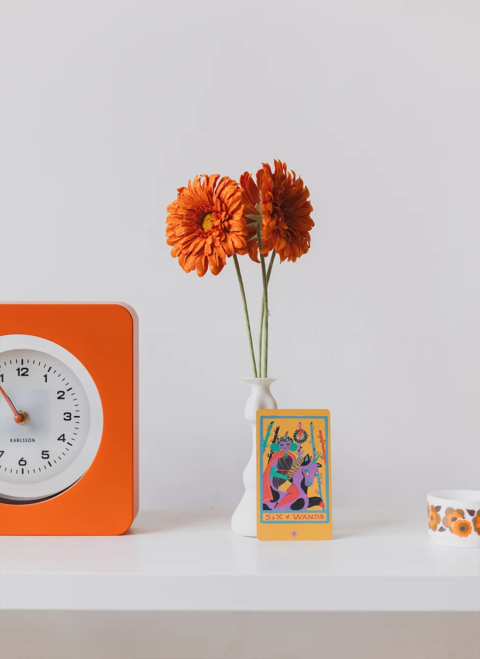 Horloge orange avec une carte de tarot adossée à un vase avec une fleur