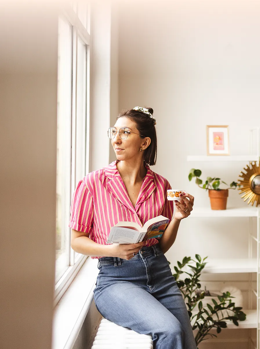 Marjorie, tarologue, assise près d’une fenêtre avec un livre et un café, dans un moment calme et introspectif